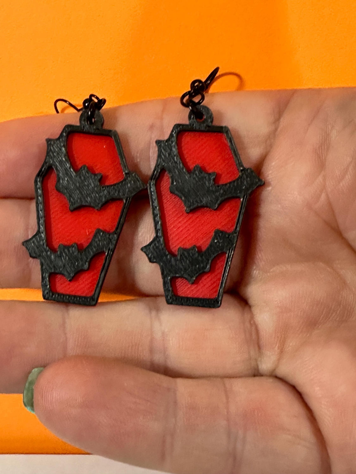 Red coffin dangle earrings with black bat cutouts on black hooks, held in hand