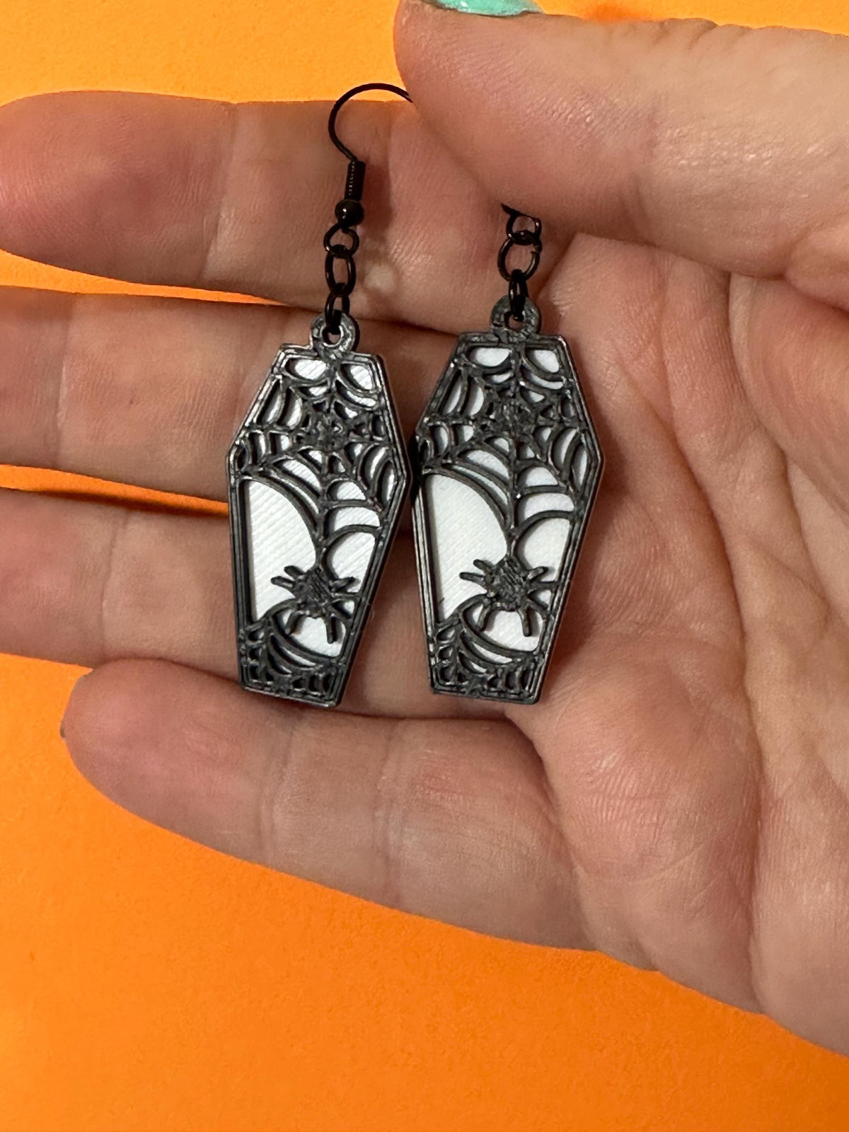 Black spiderweb coffin dangle earrings with spider detail, translucent backing, held in hand