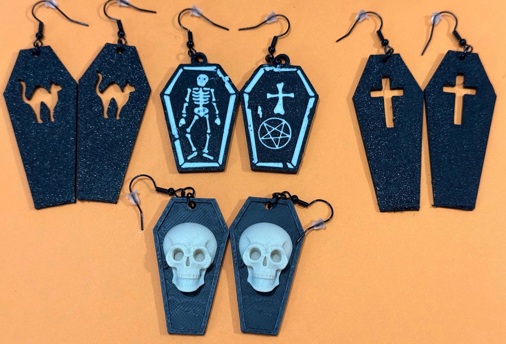 Five pairs of black coffin earrings on orange: cat, skeleton, pentagram, cross, skull designs