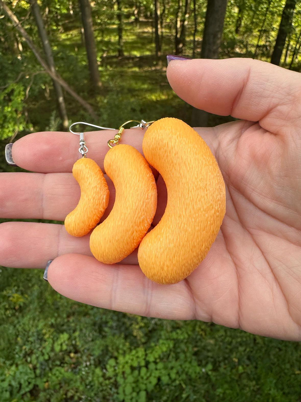 Hand holding three sizes of 3D printed cheese puff earrings, small medium and large, outdoors with trees behind