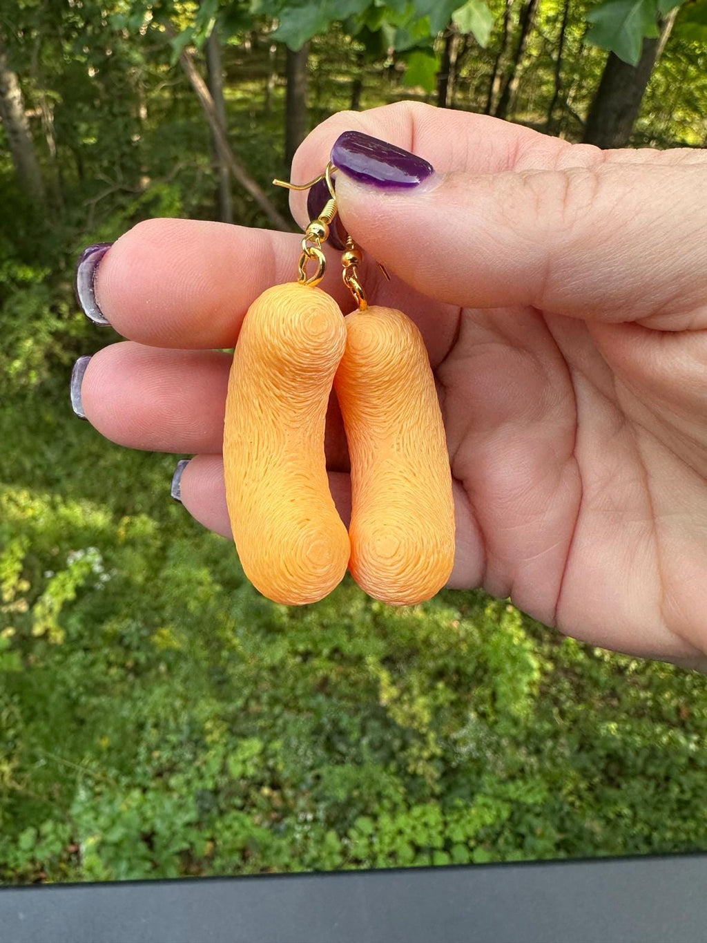 Two medium 3D printed cheese puff earrings with gold hooks pinched between fingers outdoors