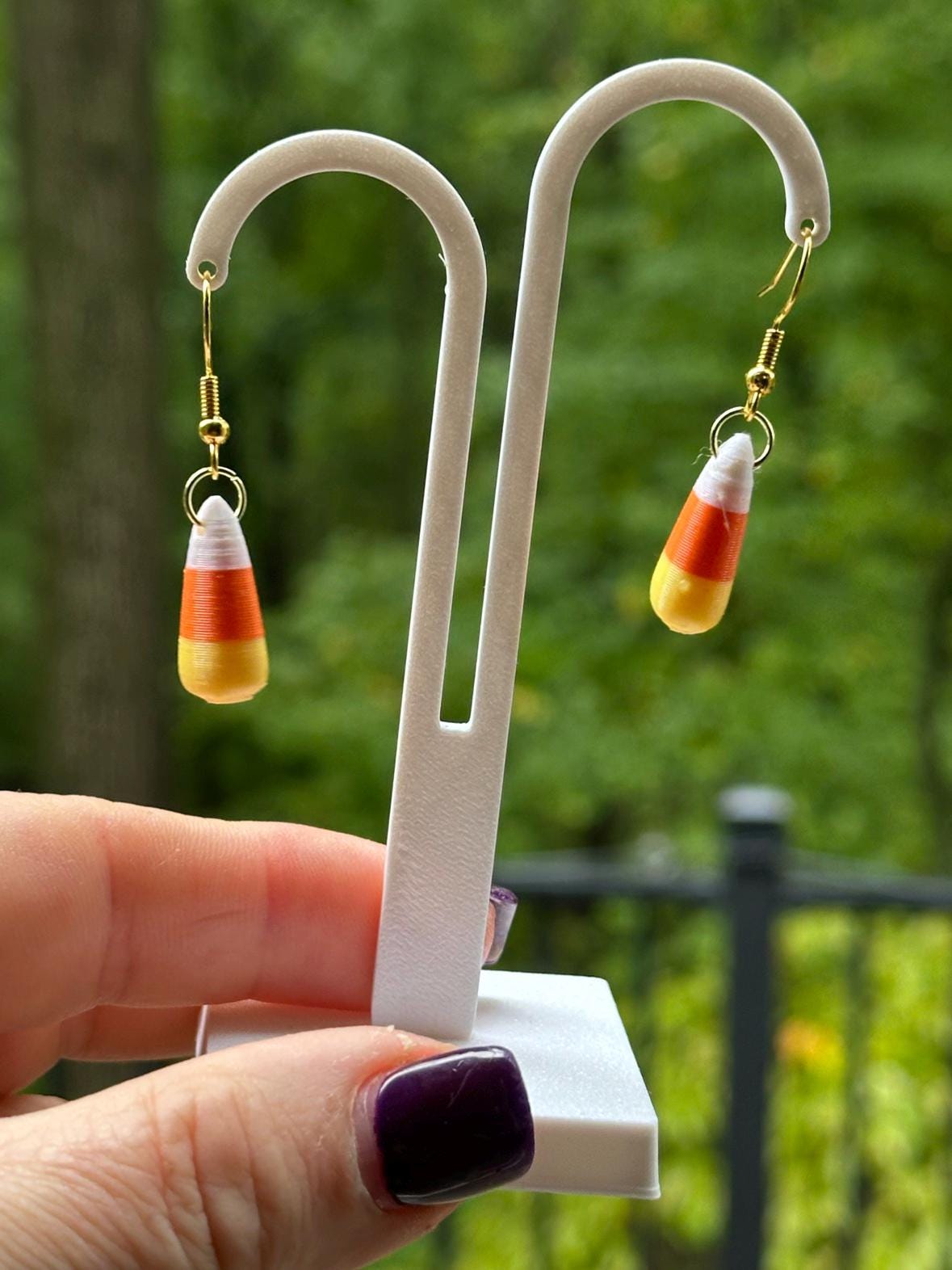 Pair of 3D printed candy corn earrings on display stand with green trees behind